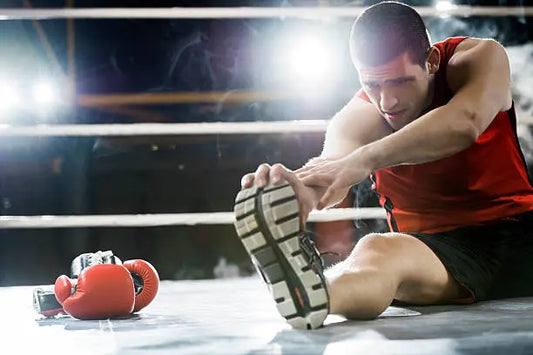Male boxer in a red tank top performing a hamstring stretch in the boxing ring, with gloves placed beside him before training.