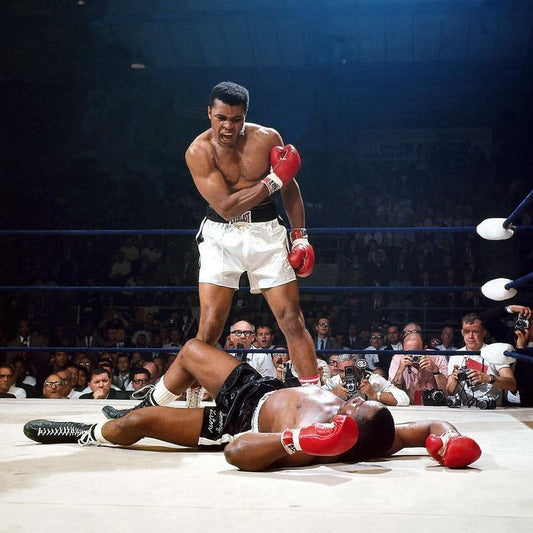 Muhammad Ali stands over a fallen Sonny Liston after a knockout, posing confidently in the boxing ring as the crowd watches the iconic moment.