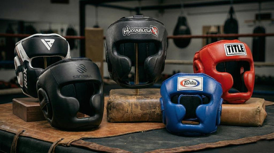 A professional display of beginner-friendly boxing headgear featuring the Hayabusa T3, TITLE Gel World, Fairtex HG3, Sanabul Essential, and RDX Maya Hide models in a boxing gym setting.